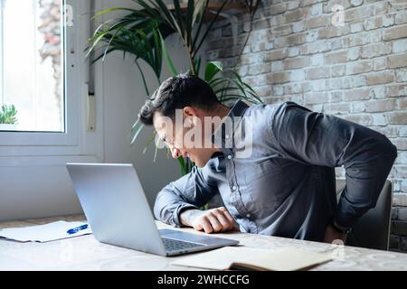 Unhappy Caucasian man sit at desk in home office work on laptop suffer from lower back muscle strain or spasm. Unwell unhealthy male have backache from incorrect posture. Sedentary life concept. Stock Photo