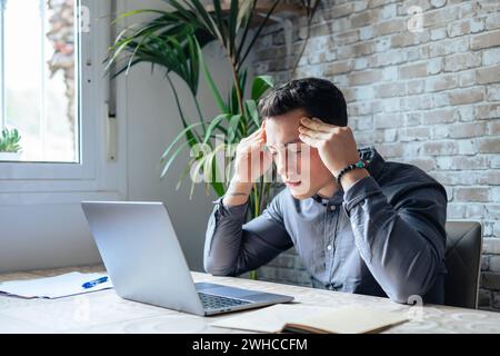 Stressed tired man in pain having strong terrible headache attack after computer laptop work, fatigued exhausted guy suffering from chronic migraine massaging temples to relieve head ache tension Stock Photo