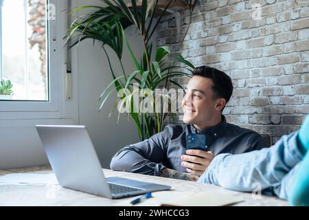 Satisfied teenager happy talking on phone at work with laptop in office, raises hands and puts feet up on table, relaxing after hard working day in expectation of weekend leave, relaxed workday, no stress Stock Photo