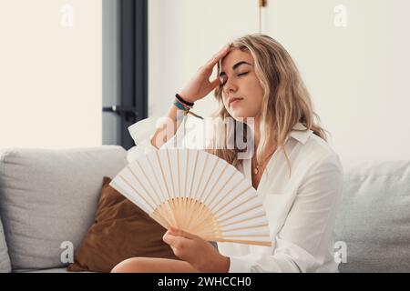 Overheated female sitting on couch in living room at hot summer weather day feeling discomfort suffers from heat waving white fan to cool herself, girl sweating dwelling without air conditioner concept Stock Photo
