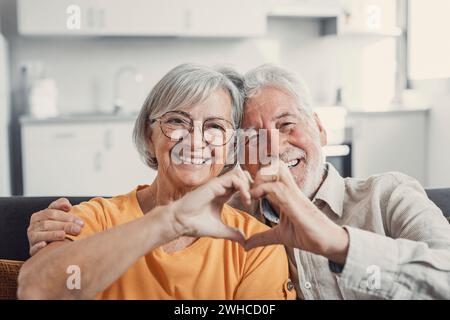 Close up portrait happy sincere middle aged elderly retired family couple making heart gesture with fingers, showing love or demonstrating sincere feelings together indoors, looking at camera. Stock Photo