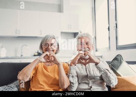Close up portrait happy sincere middle aged elderly retired family couple making heart gesture with fingers, showing love or demonstrating sincere feelings together indoors, looking at camera. Stock Photo