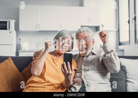 Happy older couple sit on couch staring at cellphone screen gesturing looking overjoyed, scream with joy, read fantastic news, get great commercial offer, pension raise. Success, achievement concept Stock Photo
