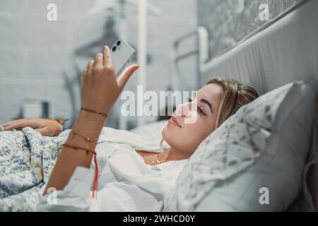 Gadget addiction. Calm smiling young woman or teenage girl lying at cozy bed holding cellphone in hands looking at screen chatting checking social network account before fall asleep or after waking up Stock Photo