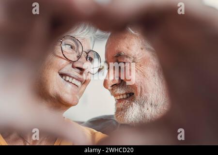 Close up portrait happy sincere middle aged elderly retired family couple making heart gesture with fingers, showing love or demonstrating sincere feelings together indoors, looking at camera. Stock Photo