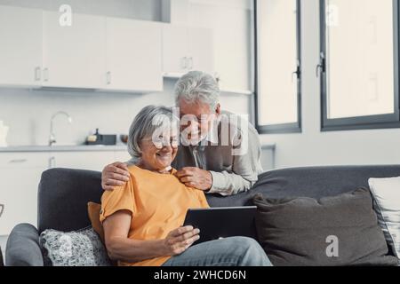 Happy older couple using computer tablet together at home, excited mature man and woman looking at mobile device screen, shopping or chatting online, sitting on cozy couch in living room Stock Photo