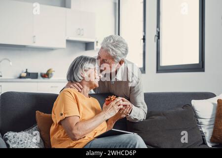 Smiling senior husband making surprise giving gift box to amazed wife, happy aged woman get unexpected present from spouse spending romantic weekend or celebrating anniversary together Stock Photo