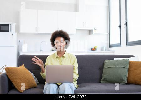 Easy learning. Smiling young mixed race lady sit on couch search information using laptop watch webinar video lesson. Positive black woman use online education service to improve skills waving Stock Photo