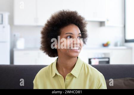 Head shot happy dreamy young African American sincere woman looking in distance, resting on cozy sofa. Inspired young pretty woman daydreaming, visualizing future, recollecting good memories indoors. Stock Photo