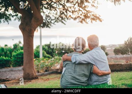 Head shot close up portrait happy grey haired middle aged woman snuggling to smiling older husband, enjoying tender moment at park. Bonding loving old family couple embracing, looking sunset. Stock Photo