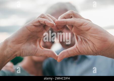 Close up portrait happy sincere middle aged elderly retired family couple making heart gesture with fingers, showing love or demonstrating sincere feelings together outdoors, looking at camera. Stock Photo