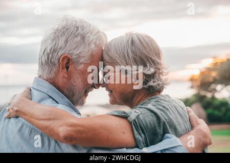 Head shot close up portrait happy grey haired middle aged woman snuggling to smiling older husband, enjoying sitting on bench at park. Bonding loving old family couple embracing, looking sunset. Stock Photo