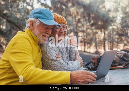 Portrait, close up of cute couple of old middle age people using computer pc outdoors sitting at a wooden table in the forest of mountain in nature with trees around them. Stock Photo