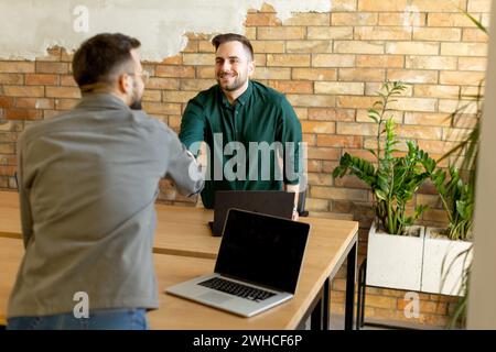 Two professionals engage in a welcoming handshake across a wooden table adorned with laptops, signaling a successful meeting or partnership in a conte Stock Photo