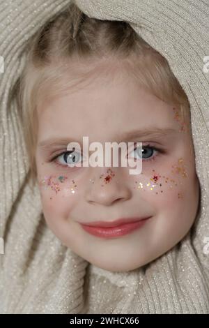 Portrait of cute little blonde girl with blue eyes and glitters on her face. Stock Photo