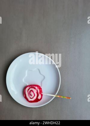 A red and white lollipop with a stick lies on a plate against a light gray background Stock Photo