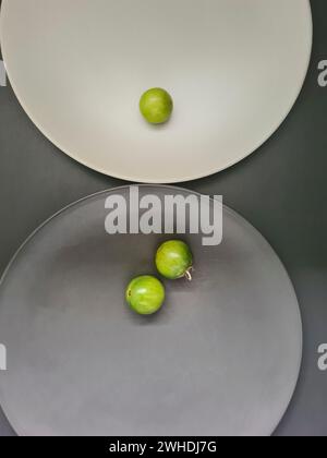 a white plate with 1 green tomato and a gray plate with 2 small tomatoes on a gray background on the theme of contrasts and minimalism Stock Photo