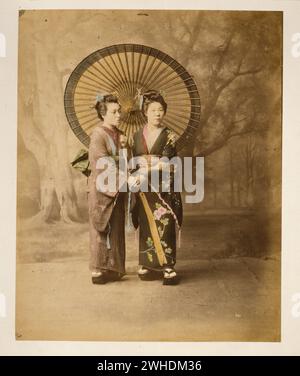 Two Japanese women in kimono, full-length studio portrait with backdrop, facing slightly right, standing with large parasol and holding hands....Japan  Hand-coloured with watercolour Photographic Print circa 1870s Stock Photo