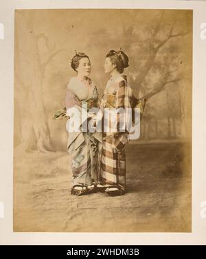 Two Japanese women in kimonos, full-length studio portrait with backdrop, standing, facing each other, holding hands- ca. 1877 ....Japan  Hand-coloured with watercolour Photographic Print circa 1870s Stock Photo