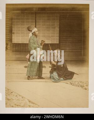 Samurai or retainer kneeling before government official or daimyo-  Photograph shows a man, full-length, standing, facing right, and a man wearing a sword kneeling before him, facing left, holding another sword as though offering it to the man standing in front of him. Exterior view of building in the background.....Japan  Hand-coloured with watercolour Photographic Print circa 1870s Stock Photo