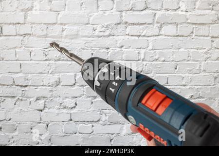 Man's hand holding Bosch cordless drill, with inserted masonry drill bit, background white brick wall, Stock Photo