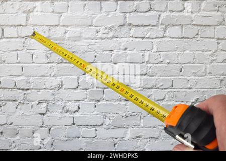 Man's hand holding a yellow measuring tape with millimeter and centimeter divisions, measuring tool, white brick wall in the background, Stock Photo