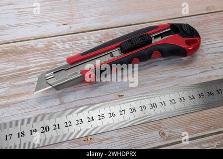 Cutter knife with 18 mm snap-off blade, plastic with rubberized black grip zone, stainless steel ruler, wooden background, Stock Photo