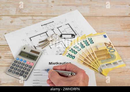 Man's hand with ballpoint pen, contract, rental deposit, bunch of keys with black key ring, without inscription, calculator, euro bills, floor plan, wooden background, Stock Photo