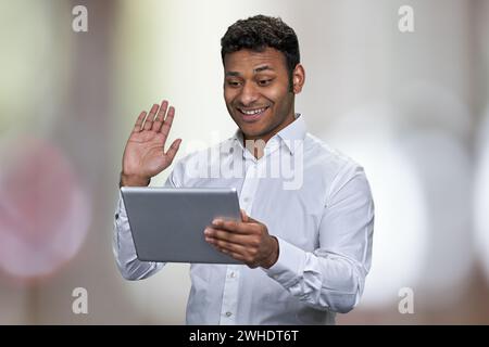 Happy young businessman waving while having video call on digital tablet. Abstract bokeh background. Stock Photo