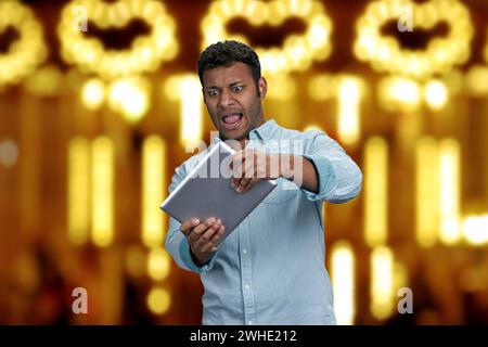 Emotional funny young man palying game on digital tablet. Golden bokeh lights background. Stock Photo