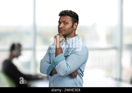 Thoughtful young man holding hand under chin. Office interior blur background. Young man with suspicious expression. Stock Photo