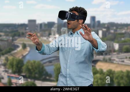 Young shocked man wearing vr glasses outdoor. Blur city skyscrapers in the background. Scared surprised man wearing vr headset. Stock Photo
