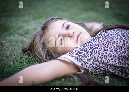 Hermosa muchacha rubia con gracia mientras disfruta de la naturaleza en un escenario al aire libre lleno de verdor y luz y recostada en el césped Stock Photo