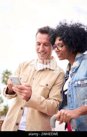 Concentrated multiracial mature tourist couple smiling standing looking and using mobile phone. Stock Photo
