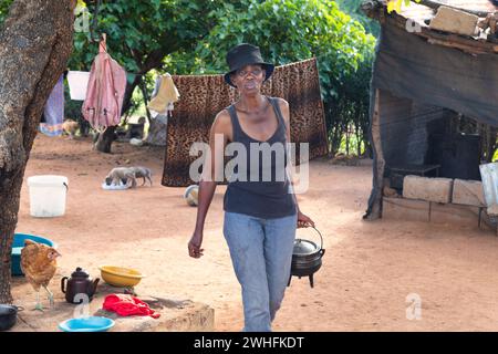 village african woman walking in the yard with a three legged pot in her hand, in front of the outdoors kitchen Stock Photo