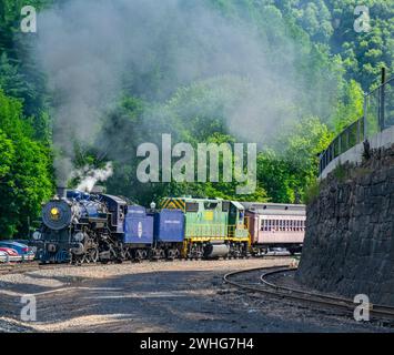 View of a Restored Steam Passenger Train Blowing Smoke and Steam Traveling Pulling Out of a Station on a Sunny Day Stock Photo