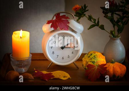 Fall back, time change on a white alarm clock after daylight saving time, candle, leaves and pumpkins as autumn decoration Stock Photo