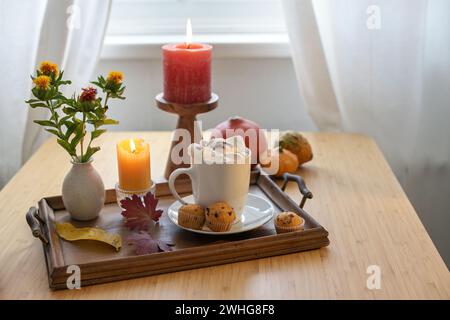 Hot chocolate drink with melting marshmallows, muffins, candles, flowers and autumn pumpkins on a table at the window, copy spac Stock Photo