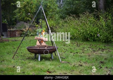 Stacked firewood in a fire bowl burns with flames and smokes under a tripod grill in a garden, preparation for a barbecue party, Stock Photo