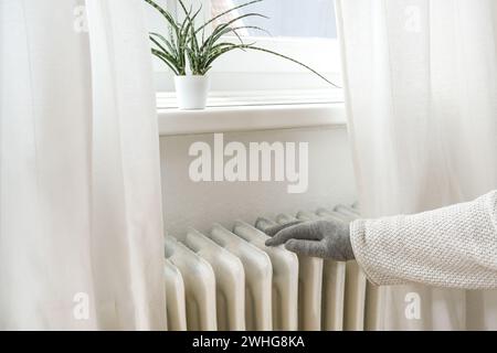Woman with warm clothes and gloves puts her hand on an old heater to feel the temperature, saving energy due to inflation and ri Stock Photo
