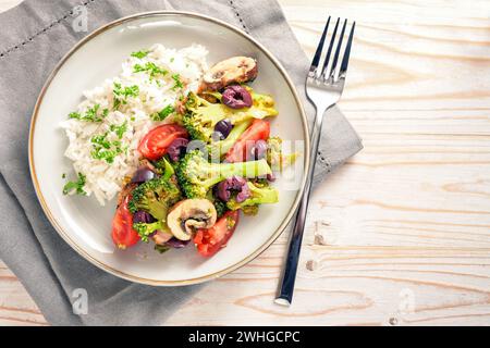 Fresh vegetables like broccoli, tomatoes and olives with rice on a gray plate and on a bright wooden table with napkin and fork, Stock Photo
