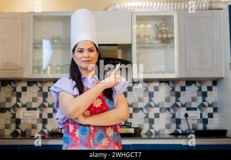 Beautiful indian woman dressed in cook uniform wearing a apron, chef hat standing in kitchen gesturing, pointing with hands. Marketing & Promotion Con Stock Photo