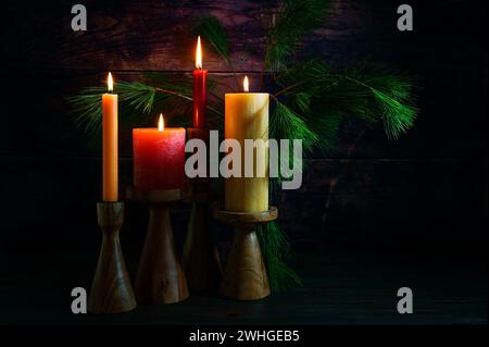 Advent arrangement with four candles in red and yellow on wooden holders and pine branches against a very dark background from r Stock Photo