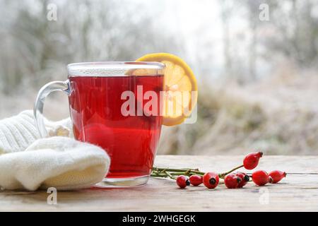 Hot red rose hip tea with a lemon slice in a glass mug on a rustic wooden table against a frosty winter landscape, copy space Stock Photo