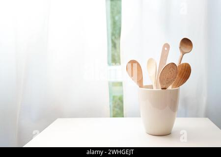 Ceramic pot with wooden cooking spoons on a table in front of the kitchen window with white curtains, copy space Stock Photo