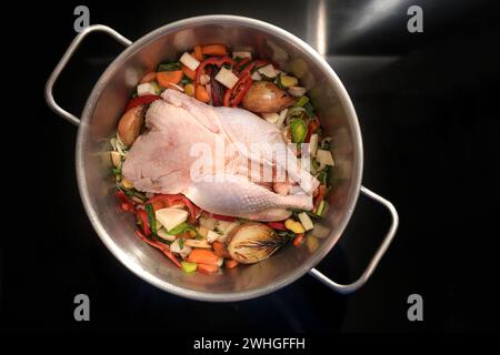 Ingredients for a healthy soup with a whole chicken and vegetables in a stainless-steel pot on the black stove, cooking for the Stock Photo