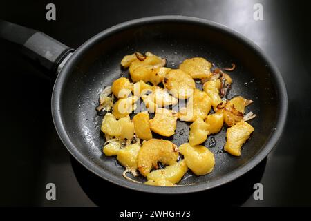 Potato slices and onion are roasted in a black frying pan on the stove, in Germany called Bratkartoffeln, food and cooking theme Stock Photo