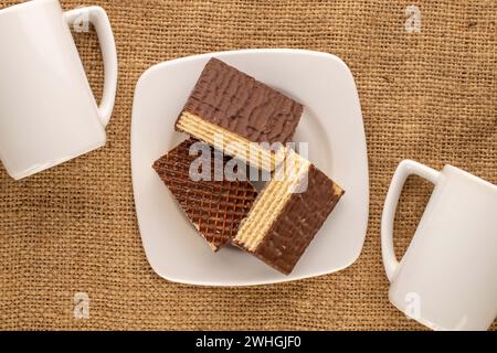 Several sweet chocolate wafers with two cups on jute cloth, macro, top view. Stock Photo