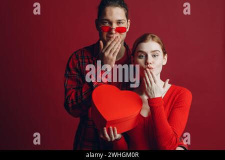 Young man gives his girlfriend a heart-shaped box in Valentine's day on red background studio Stock Photo