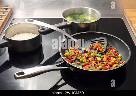 Diced vegetables are stir-fried with spices in a frying pan, pots with rice and broccoli behind them on the black stovetop, Asia Stock Photo